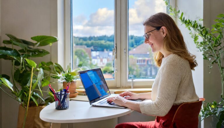 Hybrid workplace, woman working with laptop in home office by the window.